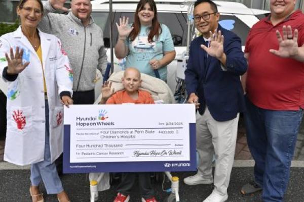 Five people hold up one of their hands toward the camera as they pose for a photo with a check from Hyundai Hope on Wheels for $400,000. A car is in the background, and behind that is Penn State Health Children’s Hospital.