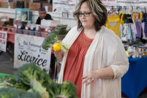 A woman holds a head of broccoli and an orange while looking at other produce at a farmer’s market. She is wearing a sweater and has glasses.