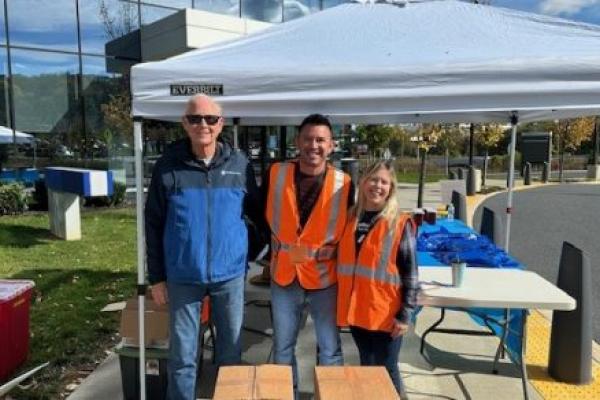 Three people wearing orange safety vests stand smiling under a white canopy tent outdoors on a sunny day.