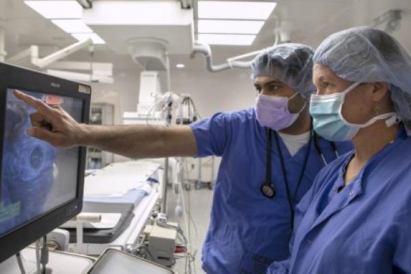 Dr. Vasudev Virparia, left, points at an intravascular ultrasound image on a monitor while Norah Martin, a registered cardiovascular invasive specialist at Penn State Health Lancaster Medical Center, watches. They are both wearing scrubs, face masks and surgical caps. They are standing in a cardiac catheterization lab.