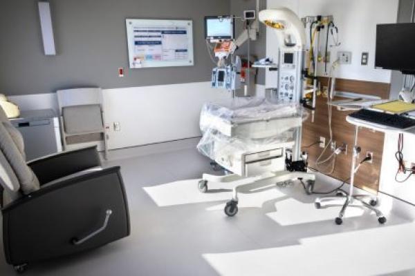 Bright, private neonatal intensive care unit (NICU) room at Penn State Health Lancaster Medical Center featuring an infant warmer with monitoring equipment, IV pumps, overhead exam light, bedside computer workstation, and a reclining chair for parents.