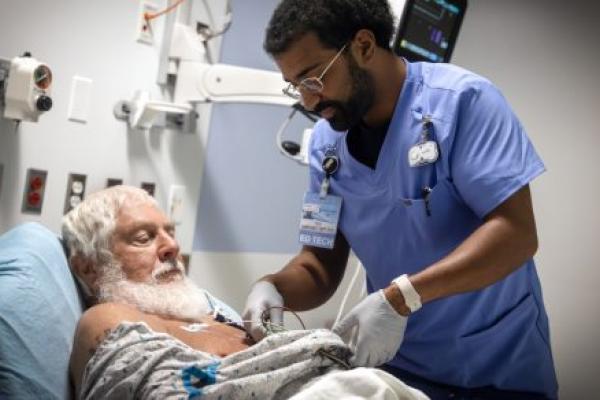 An ED technician tends to a patient laying in a bed. Various medical equipment is mounted to the wall in the background.