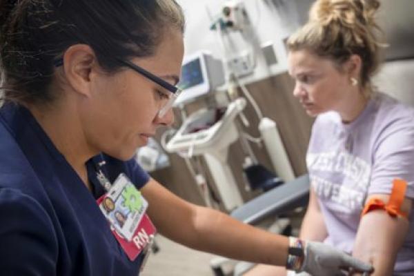 A nurse wearing navy scrubs and glasses draws blood from a patient seated in a medical chair.