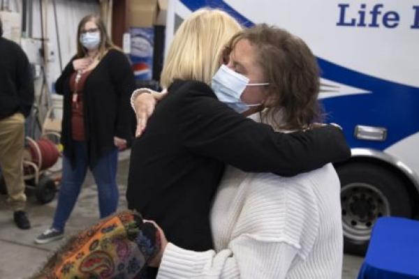 Kate Warnagiris, who has short hair and wears a bulky sweater and mask, embraces Dr. Monica Corsetti, shown from the back, who is wearing a jacket and has short hair. A Life Lion ambulance and crew members are standing are in the background.