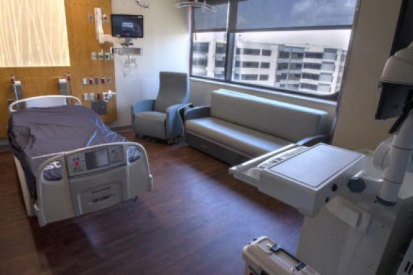 A hospital patient room with a bed, chair and sofa, as well as a headwall with various equipment and ports. A computer on a portable stand is in the foreground at right.