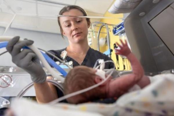 A respiratory therapist wearing scrubs and gloves adjusts breathing tubes for a premature infant lying in an incubator.