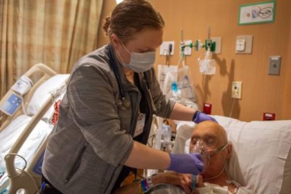 Margaret McGinnis, a respiratory therapist with Penn State Health St. Joseph, holds an oxygen mask to the face of a male patient who is lying in bed with his eyes closed. McGinnis is wearing scrubs, a sweat jacket, a lanyard with a nametag and a face mask. Behind her is another hospital bed.