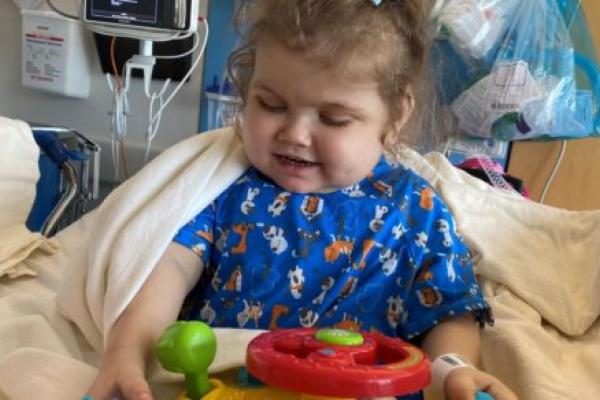 A young girl with a bow in her hair sits in a hospital bed with medical equipment behind her. She is playing with a plastic toy.