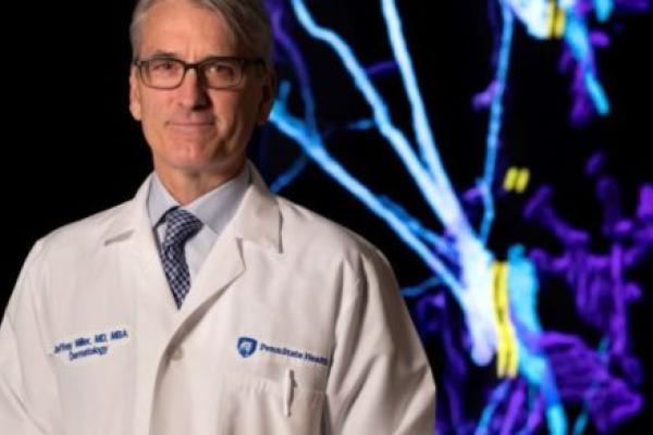 Jeffrey Miller stands in his white doctor's coat with a digital dermatology display behind him.