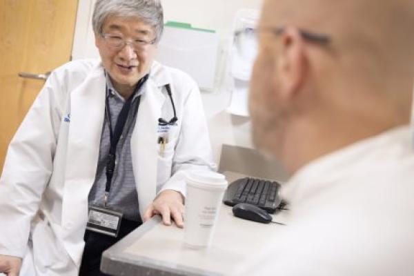 A physician wearing a white medical coat and a nametag is seated sideways in a clinic room, speaking with a patient who is in the foreground, slightly out of focus and looking away from the camera.