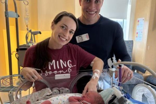 A mother and father smile in front of their infant son who is in an incubator in a hospital neonatal intensive care unit. The mother touches the baby’s hand.