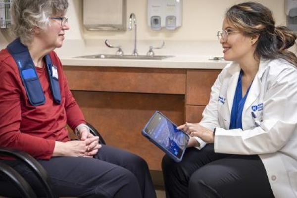 A woman patient sits across from a neurologist in a medical office. The patient has an adaptive deep brain stimulation device around her neck. The neurologist touches a tablet to adjust the device and smiles at the patient.