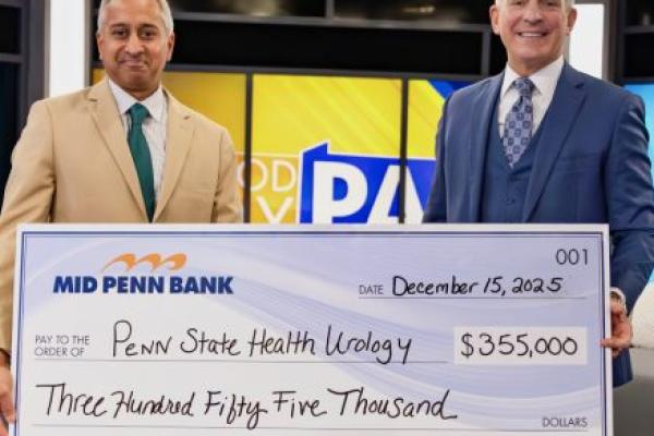 Two men in professional business attire smile as they pose for a photo with an oversized check in the amount of $355,000 made out from Mid Penn Bank to Penn State Health Urology. A TV station set is in the background.