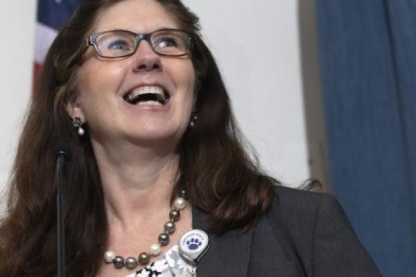 A close-up of Michele Szkolnicki speaking at a lectern. An American flag and curtain are in the background.