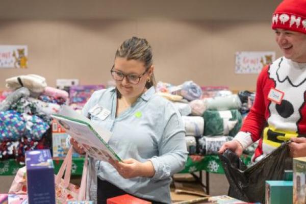 In a room full of gifts such as blankets and books, a woman looks at a children’s book. A man dressed in a seasonal “elf” outfit looks on, as he holds a bag.