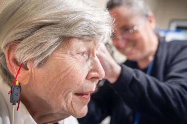 A woman is shown in profile as Dr. Jackie Price, an audiologist at Penn State Health Otolaryngology – Head and Neck Surgery, fits her with a hearing aid. A cord is hanging from the woman’s ear, and small ribbons hang from the hearing aid. Price, who is seen out of focus, is wearing scrubs, a lanyard and glasses and is smiling.