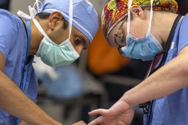 Two surgeons operate on a patient. The man on the left is wearing a face mask and scrubs. The woman on the right is wearing a colorful surgical cap, glasses, face mask and scrubs.