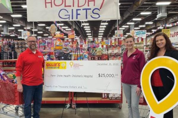 Three people pose with a check made out to Penn State Health Children’s Hospital in the amount of $25,000. A large sign reading “Holiday Gifts” is hanging above them and store merchandise is in the background.