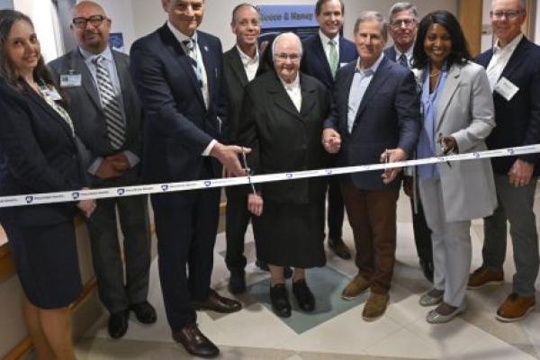 Ten people wearing professional attire pose for a photo in a hospital lobby area as they prepare to cut a ribbon with the “Penn State Health” logo.