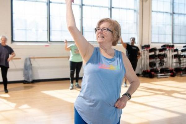 Patricia Creque puts her right arm in the air as she leads a dance class. She is wearing glasses, a T-shirt and tights. Behind her are three women, racks with hand weights and large windows.