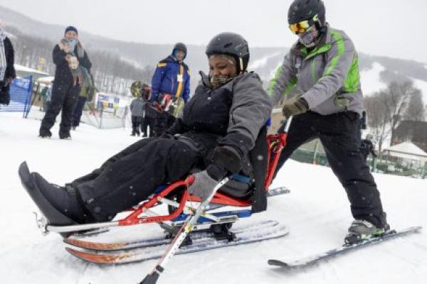 A woman is seated on an adaptive sled, while a ski instructor in winter clothing and on skis holds onto the back of the sled. They are on snow, and a few people look on from the background.