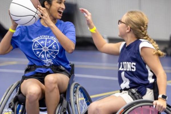 An individual in a wheelchair holds a basketball in the air and smiles. Another person to her right reaches out toward the ball.
