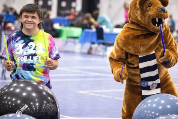 A teen boy and the Nittany Lion mascot use drumsticks to drum on exercise balls in a large gym at Penn State Health RecFest 2025. The boy is wearing a tie-dye T-shirt with the words “3.21 for Life” on it. The Nittany Lion has a scarf.