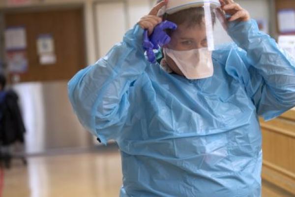 A nurse dressed in a plastic gown, mask and face shield adjusts the face shield as she walks through a hospital hallway.