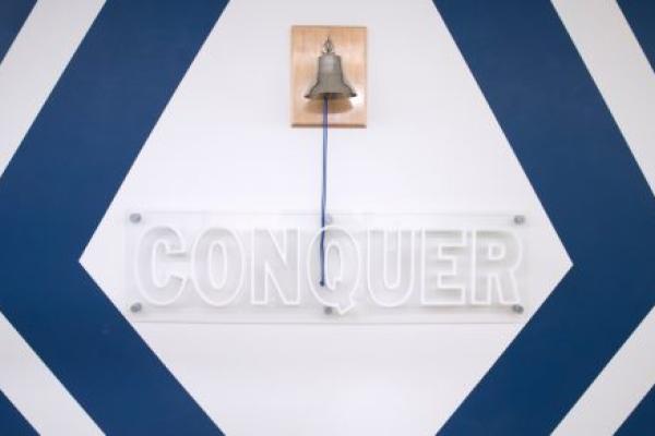 Photo shows a bell that pediatric cancer patients ring during their final cancer treatments at Penn State Health Children’s Hospital. The bell is places above a sign that says, “Conquer.”