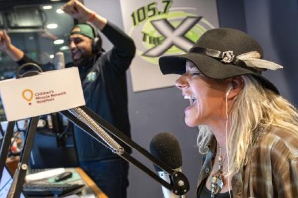 A woman wearing a black hat with feather accents laughs into a microphone while broadcasting in a radio studio. Behind her, a man wearing headphones and a backward cap raises his arms in celebration.