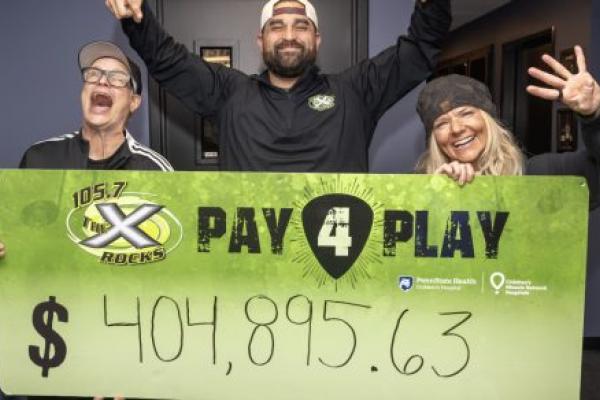 Three smiling radio hosts stand indoors holding a large green “105.7 The X Rocks Pay 4 Play” sign showing the fundraising total of $404,895.63.