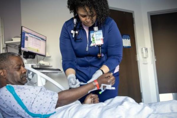 A nurse in a hospital room adjusts an IV line on a male patient lying in a bed, while monitoring equipment and a computer are visible in the background.