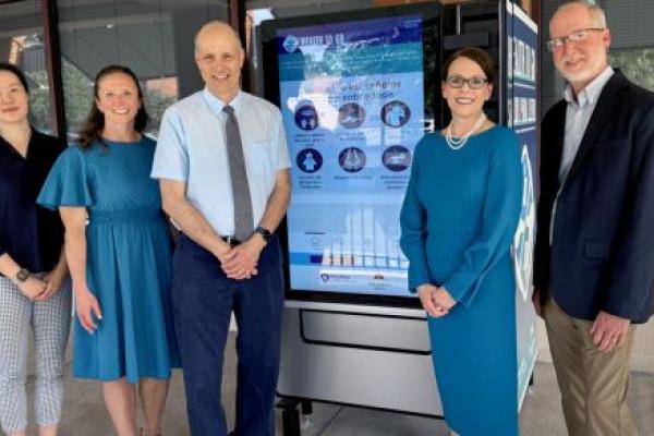A group of people stands in front of a vending machine with a digital display screen of health items.