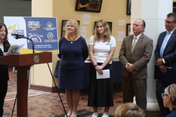 A teenage girl stands at a lectern delivering remarks. Four people stand nearby, looking on, as others – seated in front of her – listen to the comments.