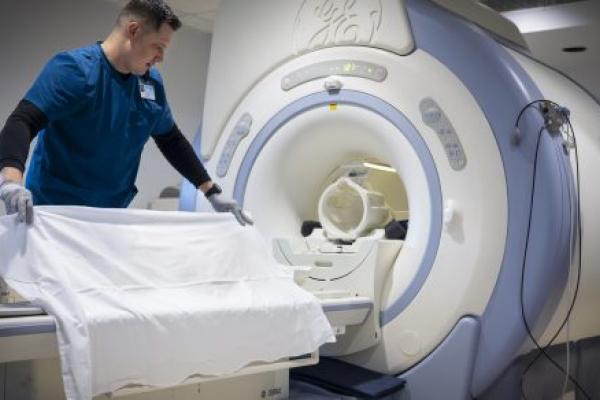A male health care worker wearing scrubs adjusts a sheet on the table of a large MRI machine in a hospital imaging suite.