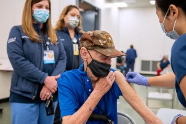 Robert Trate, wearing a mask and ball cap, rolls up his sleeve to receive a vaccination as registered nurse Judy Dee, wearing scrubs and a mask, leans toward him and cleans his bare upper arm. Physician Assistant Staci Gross Surgical Scheduler Jackelyn Ortega, wearing scrubs and masks, stand in the background.
