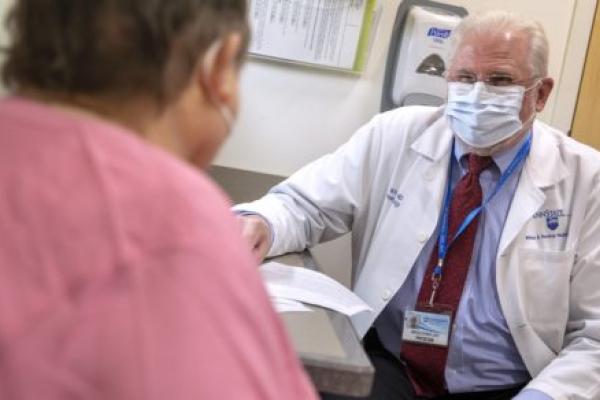 Dr. Witold Rybka, who wears a face mask, glasses and a white coat, sits in an exam room opposite a patient, who is seen from behind. A hand sanitizer dispenser and rack of papers are mounted on the wall behind him.