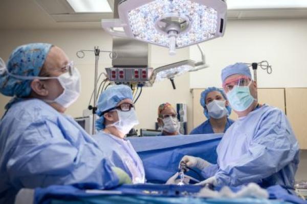 A surgical team wearing blue gowns, masks, and caps performs a procedure in a brightly lit operating room, standing around a patient draped in sterile blue sheets beneath an overhead surgical light.