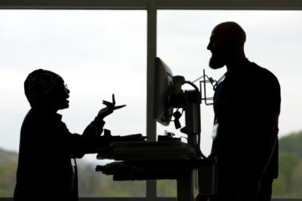 A silhouette of two employees standing and talking in front of a window at Penn State Health St. Joseph Medical Center. A computer on a cart is between the two of them.