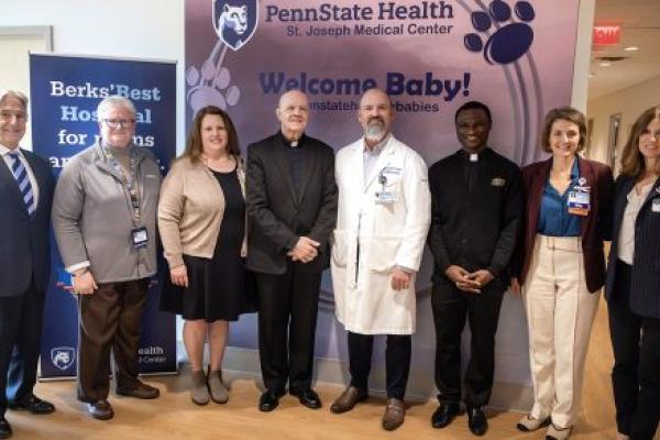 A group of eight people pose for a photo at the Breidegam Family Birthing Center at St. Joseph Medical Center. They stand in front of two banners. The one behind them reads “Penn State Health St. Joseph Medical Center” and “Welcome Baby!”