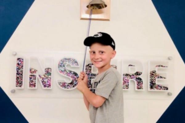A boy wearing a baseball cap faces the camera and smiles, standing in front of a bell that hangs on a wall along with the word "INSPIRE"