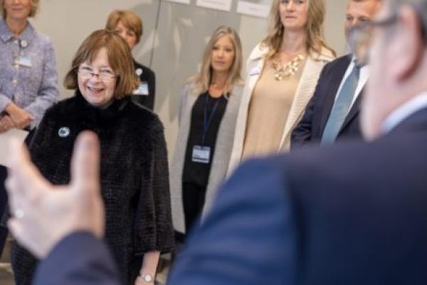 A group of people form a semi-circle around a woman who smiles as she looks at a man in a suit standing near the camera.