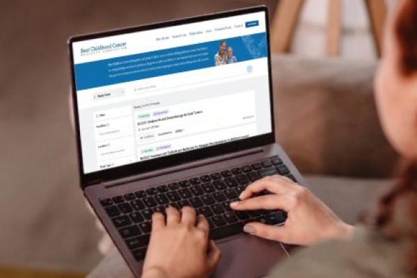 A view from over the shoulder of a woman typing on a laptop computer. On the screen is a page within the Beat Childhood Cancer Research Consortium website that displays Study Finder search results.