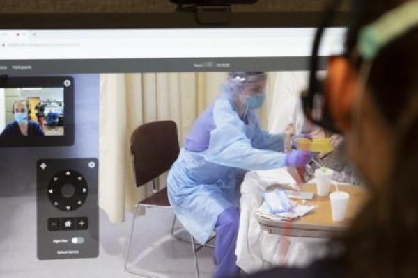 Image shows over the shoulder someone looking at a computer monitor. In one window on the monitor is the view of someone in a mask. To the right, someone in personal protective equipment works with a patient.