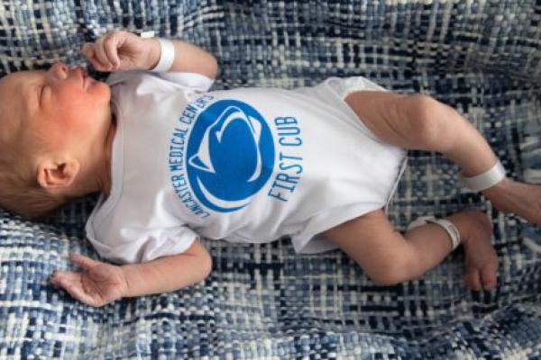 A newborn baby lays on a textured blanket. He wears a onesie with a Penn State logo and the words “Lancaster Medical Center’s First Cub.”