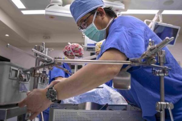 Dr. Johnny Hong, wearing surgical scrubs, works with a piece of equipment in the foreground of an operating room. Other staff members are working in the background.