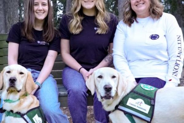 Three women sit on a wooden bench outdoors in a wooded area, smiling at the camera, each accompanied by a Golden Labrador retriever wearing a green “Canine Assistants” service vest.