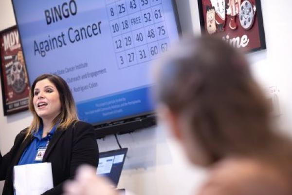 A woman stands in front of a screen with the words “BINGO Against Cancer” on it and a bingo card. She puts two fingers in the air. A woman, out of focus on the right, watches her.