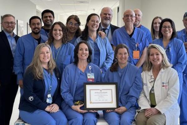Men and women, most dressed in hospital scrubs, pose in a hospital corridor. Four women seated in front, with two holding a framed certificate.