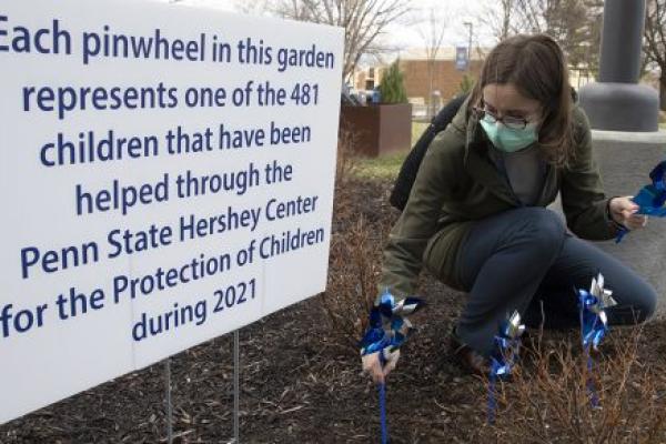 Dr. Kathryn Crowell crouches down as she puts a pinwheel in the ground in front of Penn State Health Milton S. Hershey Medical Center in 2023. A sign to her left says: “Each pinwheel in this garden represents one of the 579 children that have been helped through the Penn State Hershey Center for the Protection of Children during 2022.”